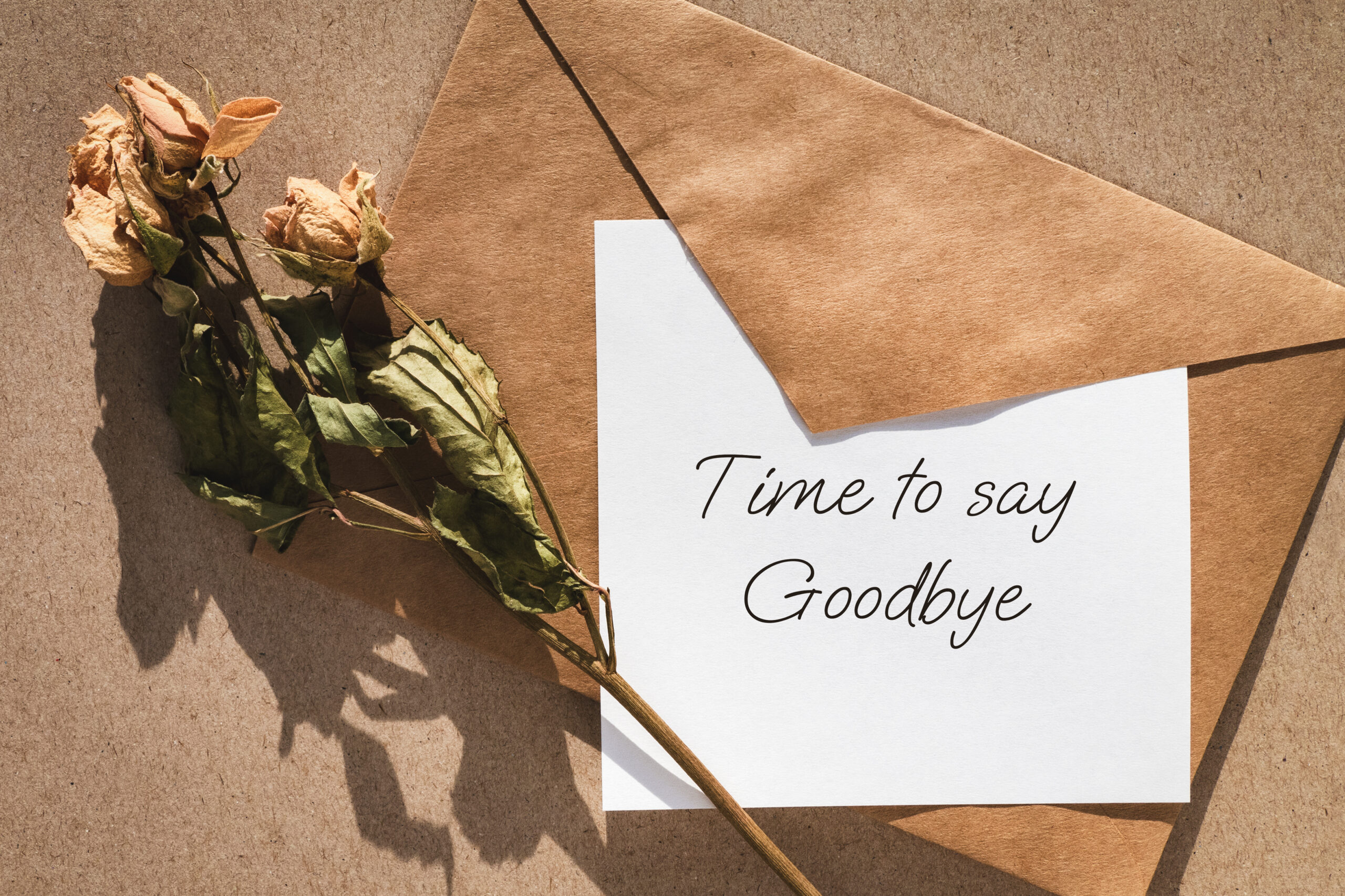 A paper that reads "Time to say goodbye" lays on top of a brown envelope. A bundle of dry roses are laid across the edge of the envelope. Sometimes falling out of love means it's time to say goodbye.