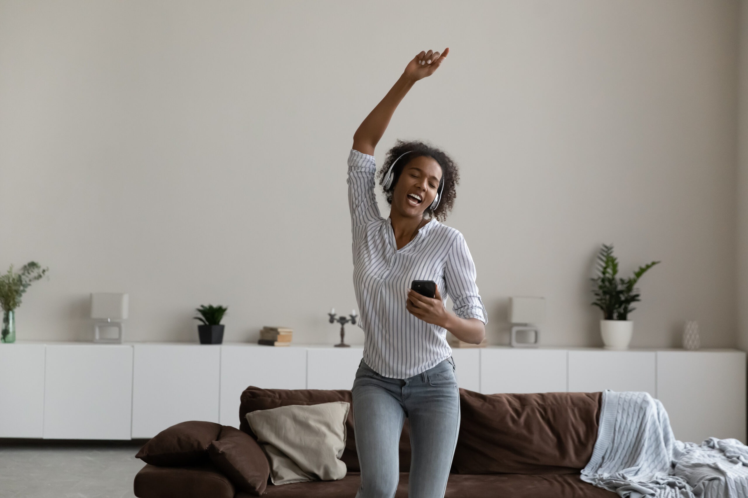 A woman is dancing with her headphones on, enjoying being alone. A brown couch is shown behind her.