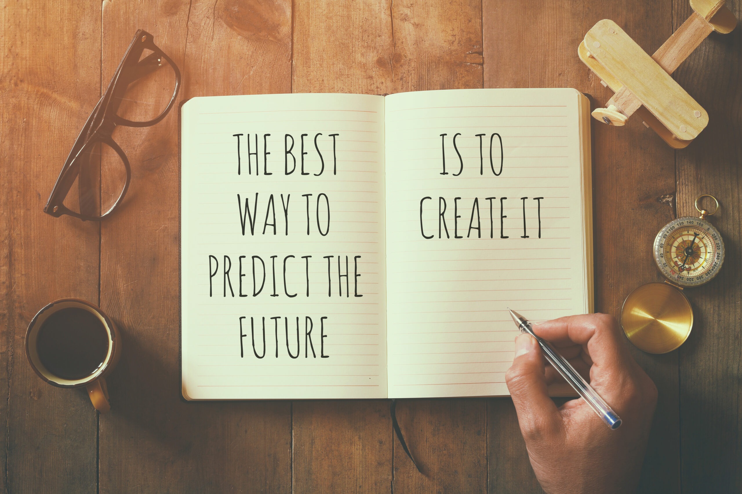 A hand holding a pen writes "The best way to predict the future is to create it" in a notebook, meaning you can't control the future, but you can learn to handle major life changes with grace.