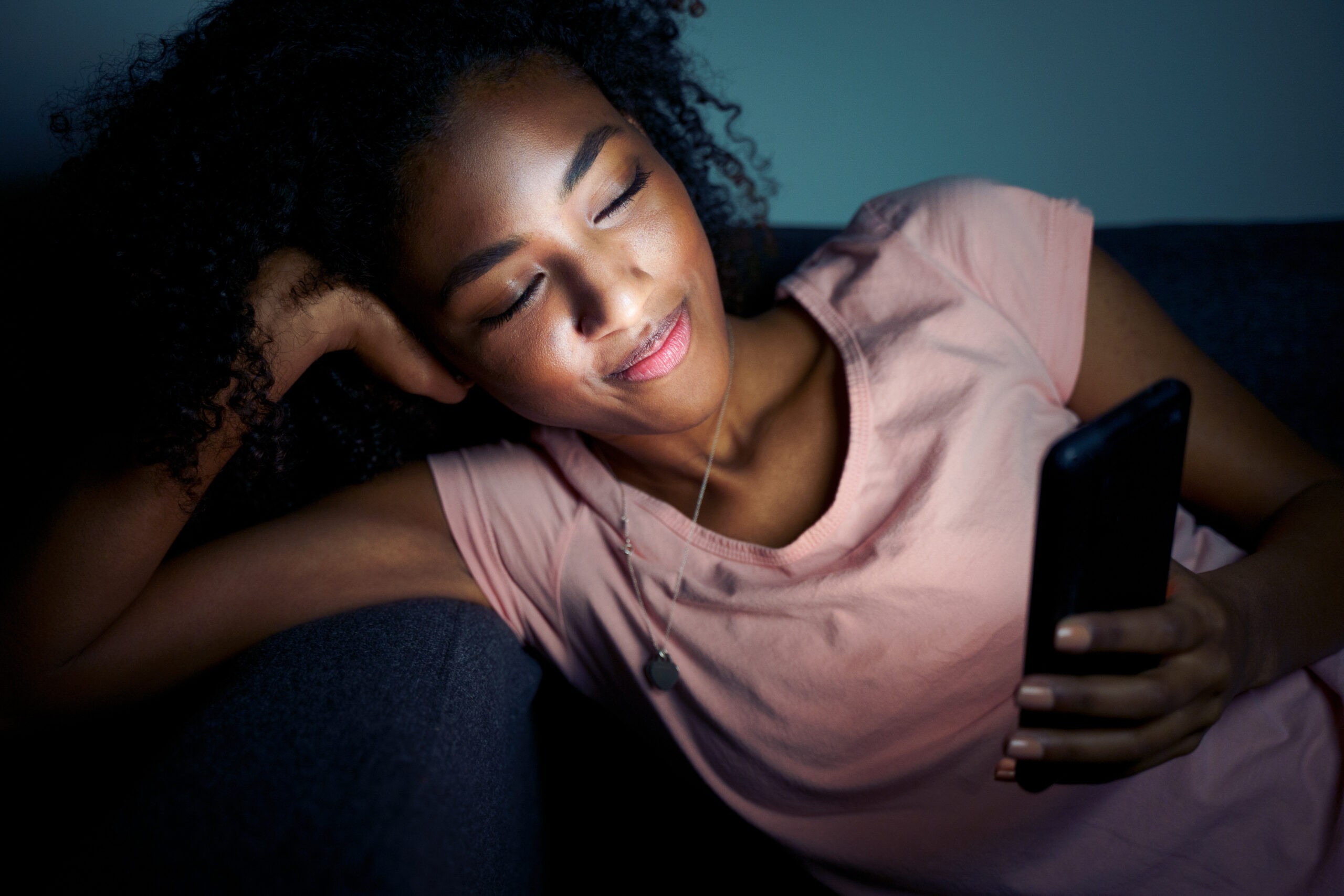 Woman finding love online smiles at her phone while leaning on her hand. It's dark and the phone light shows on her face.