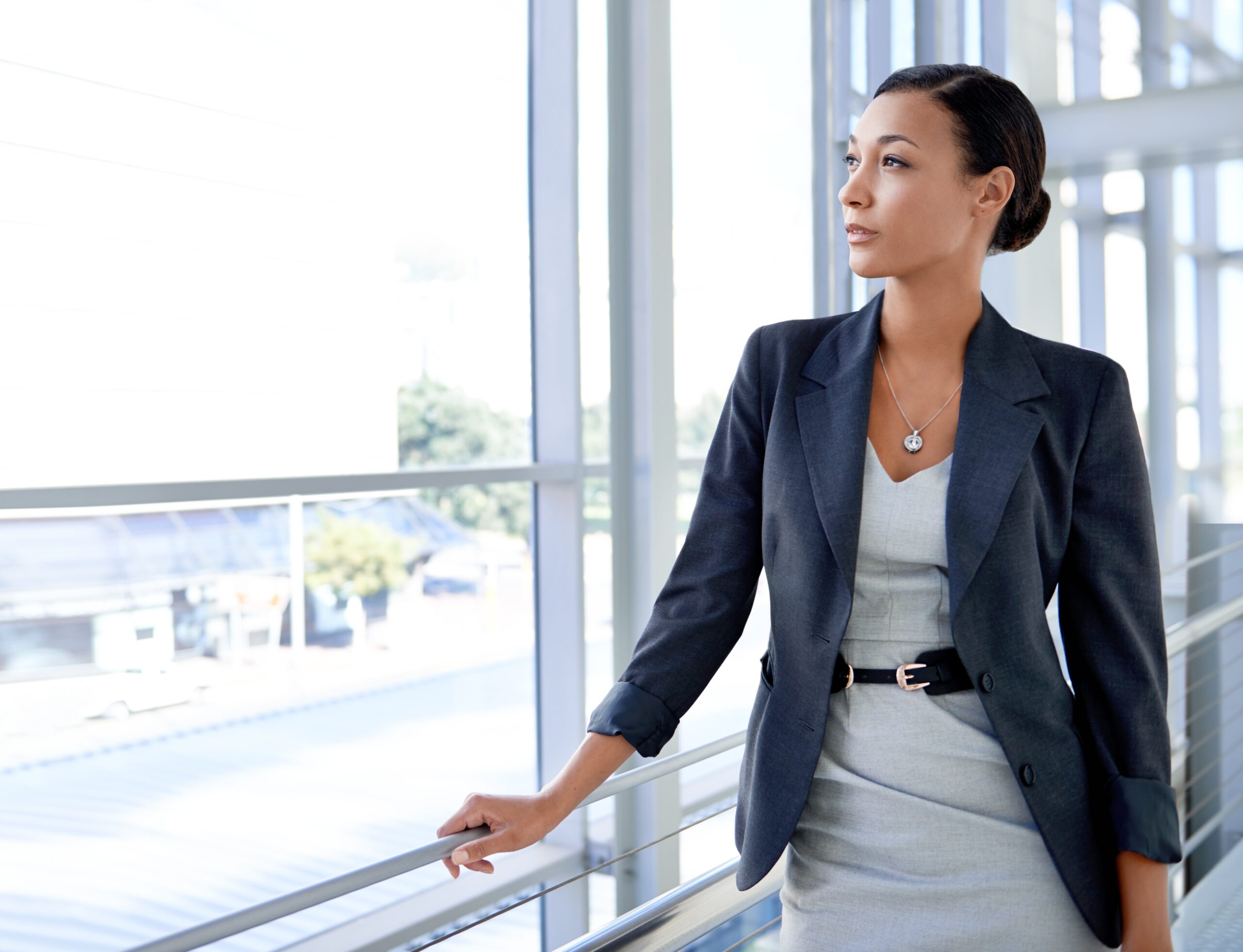 A business woman in a light blue dress and dark blue blazer stares out the window of an office building.