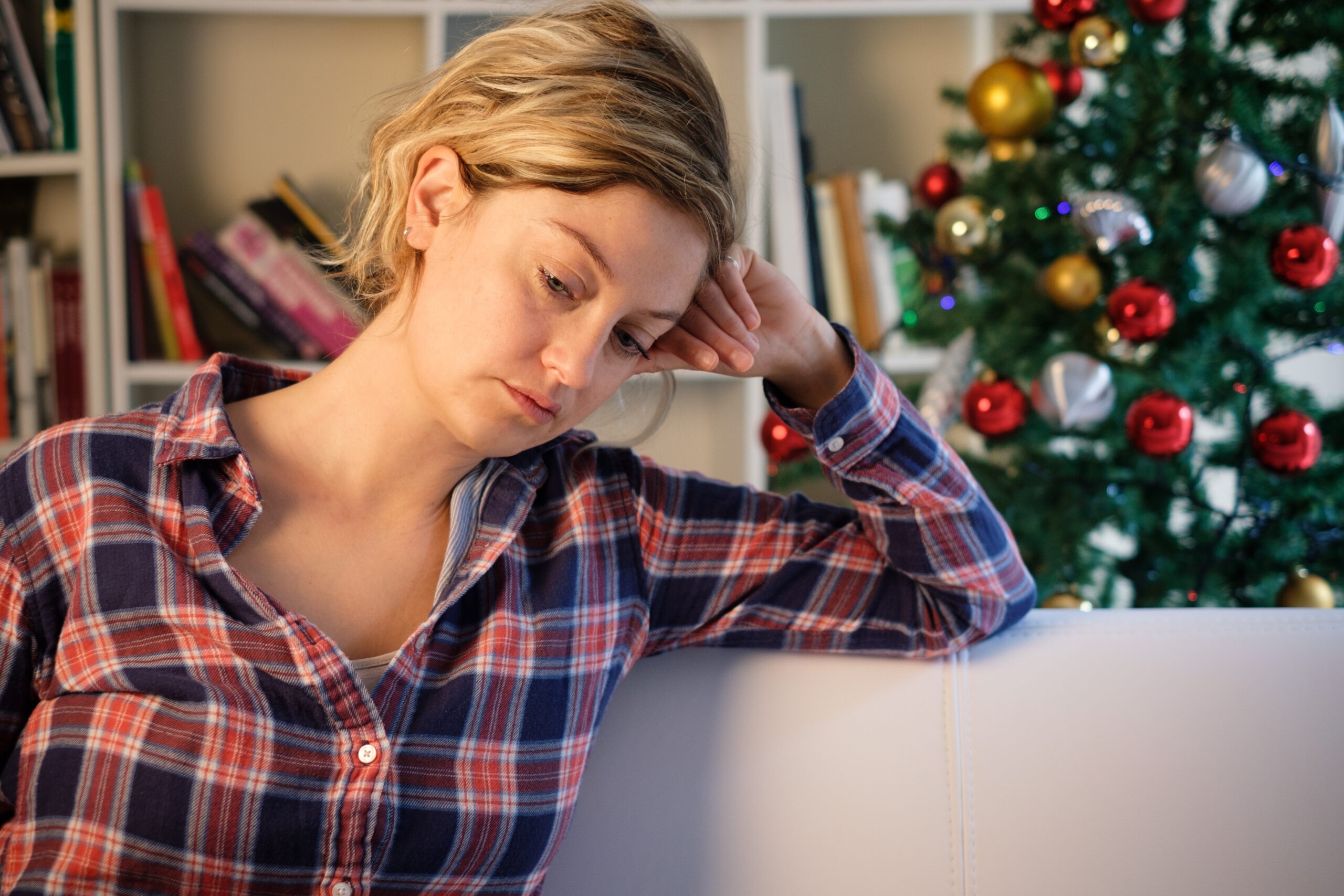 A woman in a plaid flannel sits on a couch in front of a Christmas tree. Her head leans against her hand as she stares sadly toward the ground, portraying the complexity of grieving during the holidays.