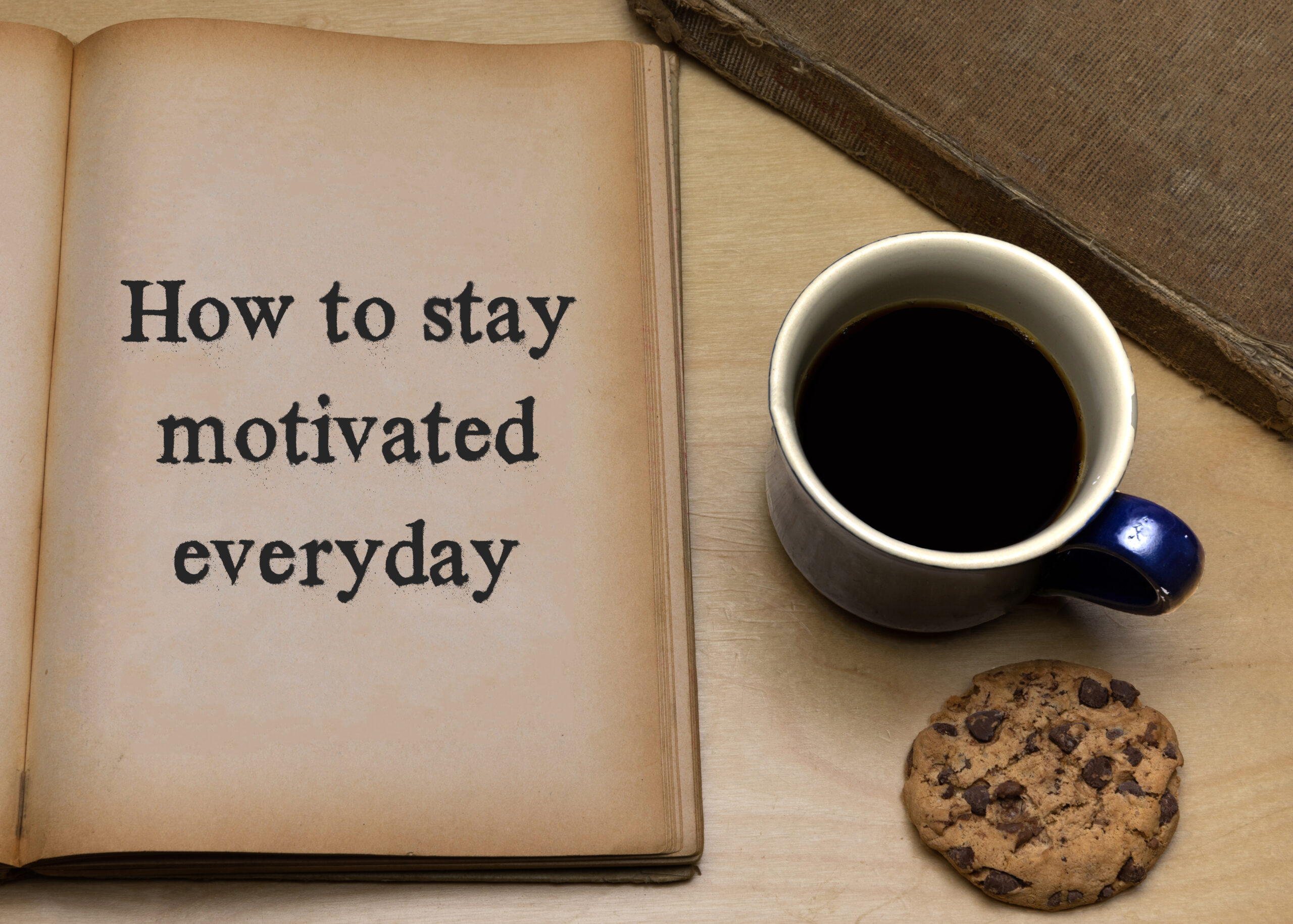 "How to stay motivated everyday" written on a notebook page. A cup of coffee and a chocolate chip cookie are next to the notebook.