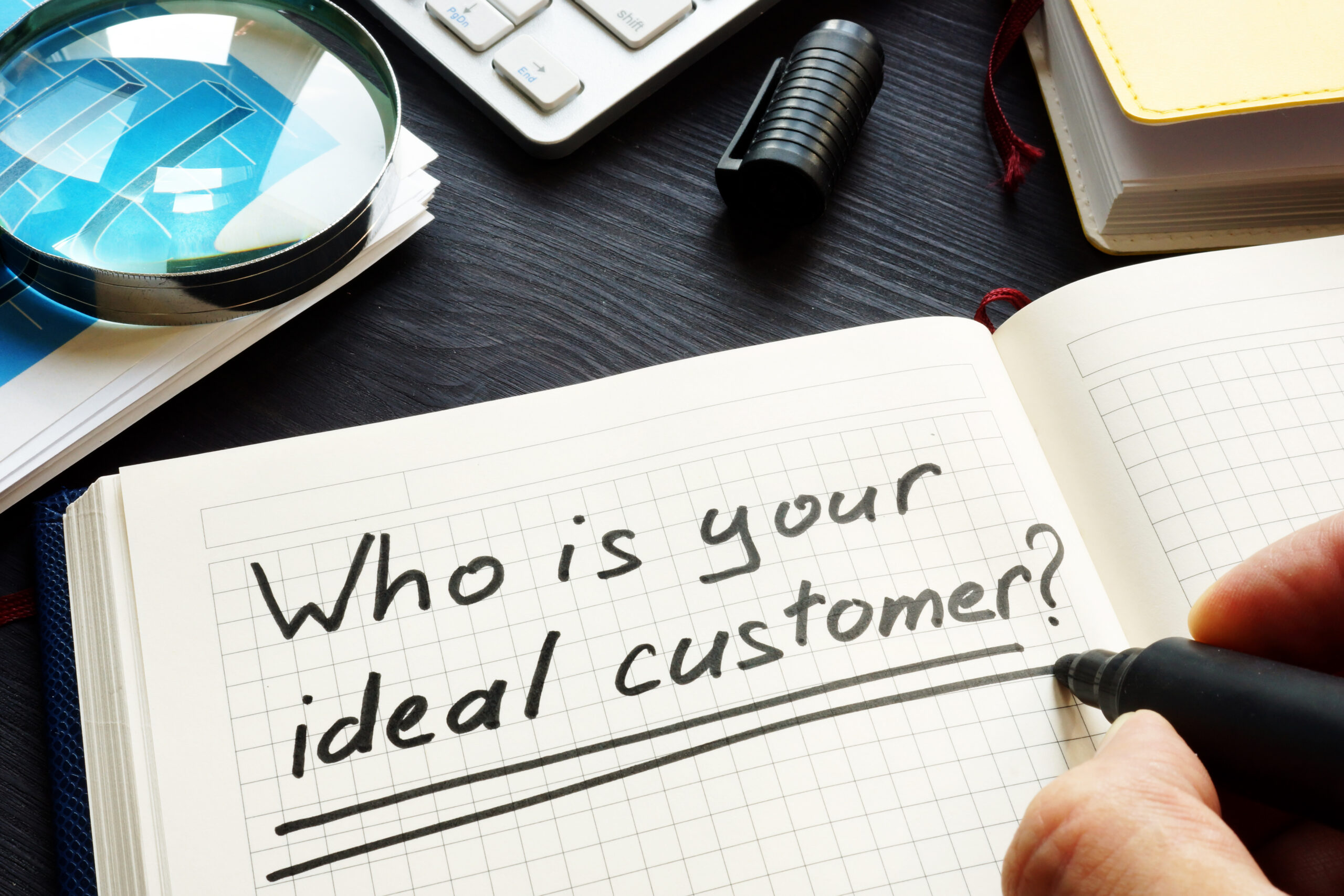 "Who is your ideal customer?" is written in a journal. The author's hand is shown holding a black marker.